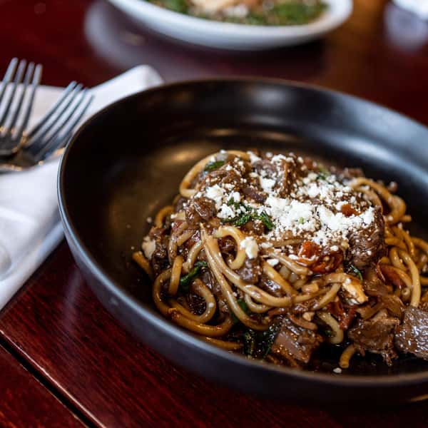 Braised beef pasta in a rich red wine sauce, topped with crumbled cheese and wilted greens in a black bowl.