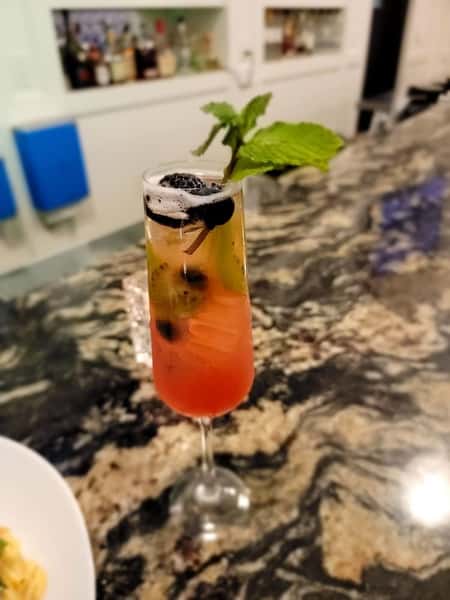 A colorful cocktail garnished with mint leaves, blackberries, and a cinnamon stick, set on a marble bar counter with a plate of food in the foreground.