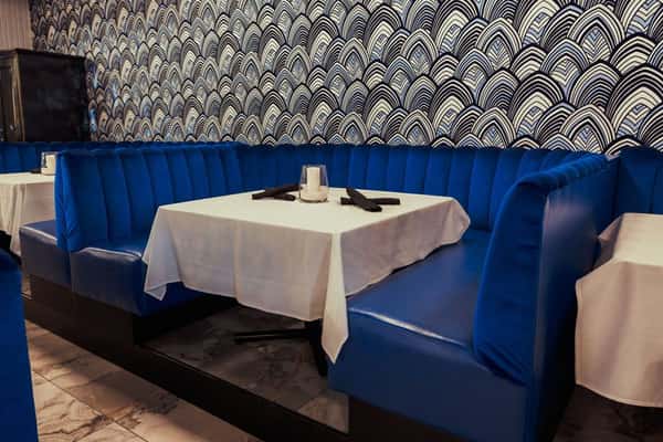 A luxurious dining area with deep blue velvet booths, white tablecloths, and a modern bar in the background with colorful lighting.