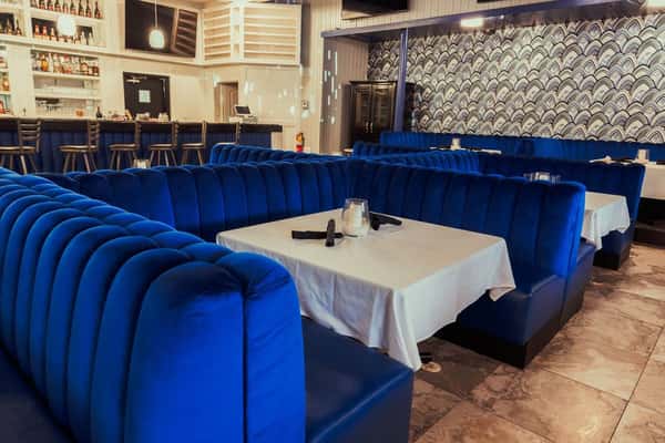 A luxurious dining area with deep blue velvet booths, white tablecloths, and a modern bar in the background with colorful lighting.
