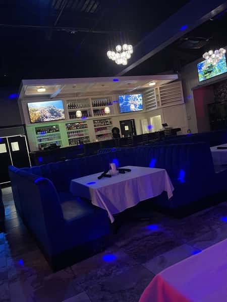 The image shows a dimly lit restaurant or bar interior with blue lighting, comfortable booths with white tablecloths, and a bar area with bottles on display and screens showing underwater visuals.