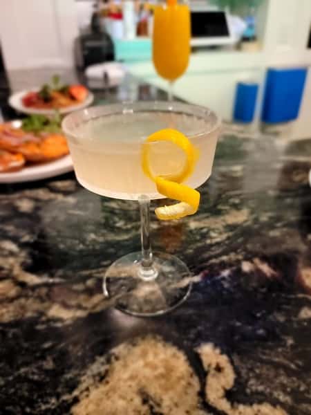 A cocktail with a twist of lemon peel, served in a coupe glass, with a background of a marble countertop and other drinks.