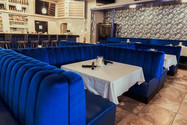 A cozy restaurant interior featuring elegant blue velvet booths with white tablecloths and a well-stocked bar in the background.
