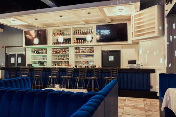 A stylish bar area with illuminated shelves displaying various bottles, a modern seating area with blue velvet booths, and two large TVs.