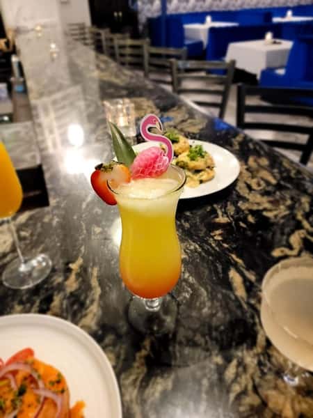 A tropical cocktail garnished with a flamingo-shaped stirrer, pineapple, and strawberry, placed on a marble bar counter with a plate of food in the background.