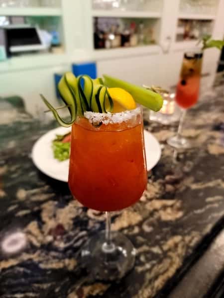 The image shows a cocktail served in a glass with cucumber slices, a lemon wedge, and a salted rim, resting on a marble bar countertop.