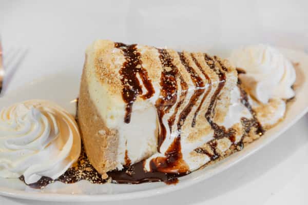 slice of s'mores cheesecake topped with chocolate drizzle and whipped cream on a dessert plate