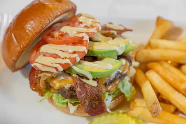 stacked avocado burger with bacon, fresh vegetables, and fries on a white plate