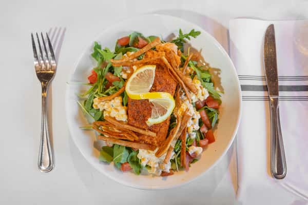 grilled salmon salad topped with street corn, fresh vegetables, and lemon wedges on a white plate
