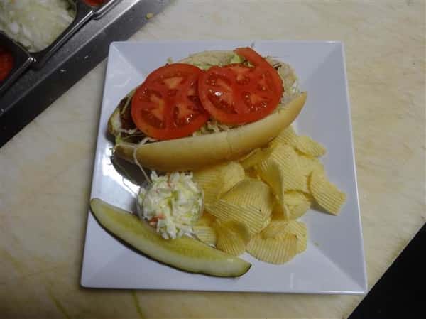hotdog on a roll topped with lettuce and tomatoes with a side of chips, coleslaw and a pickle spear