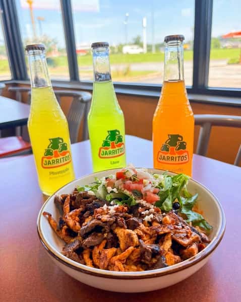 Bowl of mixed grilled meat with lettuce, pico de gallo, and cheese, with three Jarritos sodas behind it.