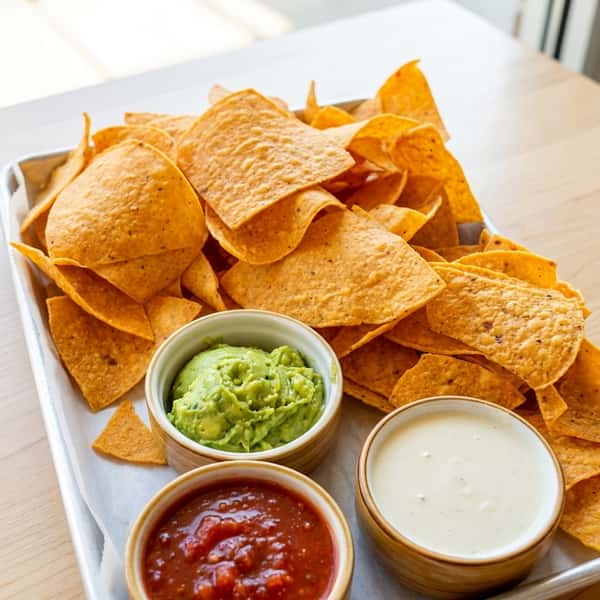 Tray of tortilla chips with bowls of guacamole, red salsa, and white queso dip.