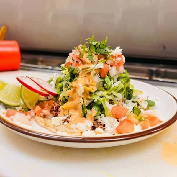 Open-faced soft taco with ground meat, lettuce, pico de gallo, cheese, sauce, and lime wedges.