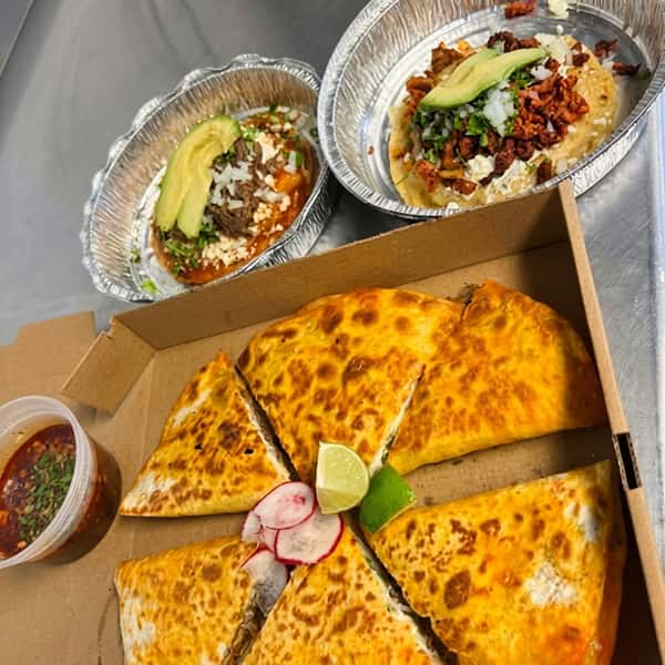 Birria pizza in a cardboard box with a side of consome, radishes, and lime. Two small quesabirrias with avocado in foil containers.