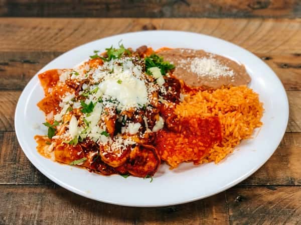 Chilaquiles rojos with shredded chicken, crumbled cheese, rice, and refried beans on a white plate.