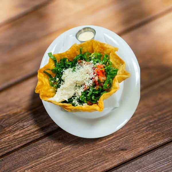 Taco salad in a fried tortilla bowl with shredded lettuce, diced tomatoes, cheese, and sour cream, on a white plate.