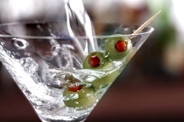 Clear martini being poured into a glass with three green olives on a skewer, creating a splash effect.