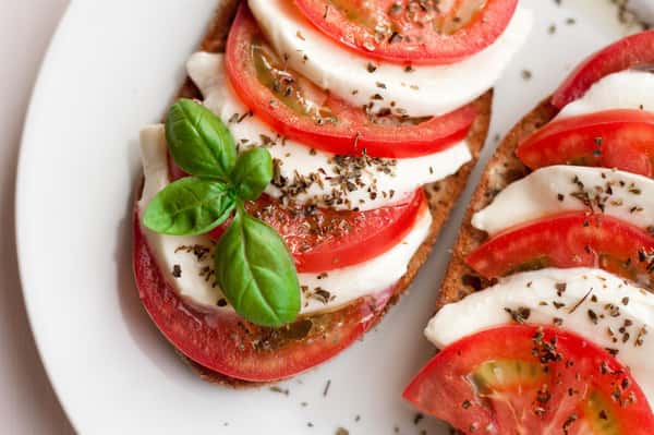 This image features a delicious Caprese-style open-faced sandwich. It's layered with fresh slices of ripe tomato and mozzarella cheese, garnished with dried herbs and a sprig of fresh basil, all served on toasted bread. A light and flavorful option, perfect as an appetizer or a refreshing snack.