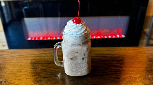 Chocolate milkshake topped with whipped cream and a cherry served in a mason jar on a wooden table with a cozy fireplace background.