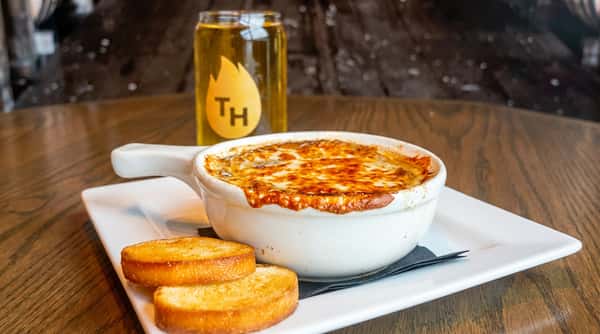 Classic French onion soup topped with melted cheese and toasted crostini served in a white crock with a glass of beer.