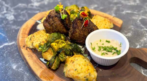 Seared steak medallions served with roasted Brussels sprouts, creamy mashed potatoes, and dipping sauce on a rustic wooden board.