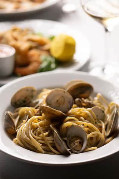 Spaghetti alle vongole served in a white bowl, garnished with parsley, with fried seafood and lemon in the blurred background.