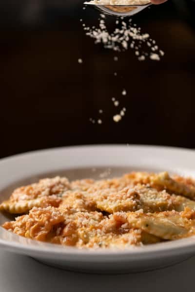 Grated cheese being sprinkled over ravioli in creamy tomato sauce on a white plate.