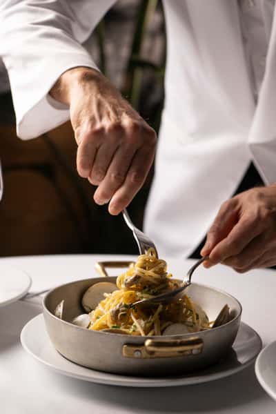 Chef in a white coat twirling spaghetti with clams in a pan using a fork and spoon on a white tablecloth.