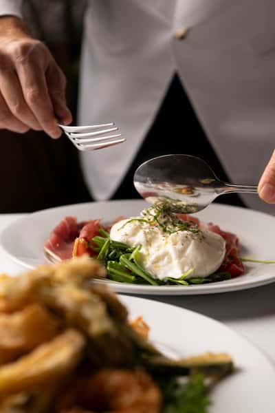 Burrata served on a bed of arugula and prosciutto, garnished with herbs and olive oil, as a person prepares to eat.