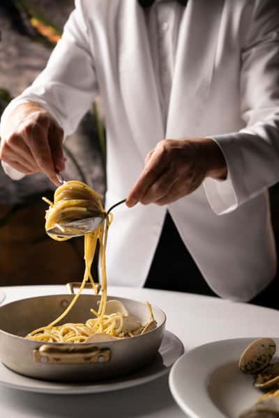 Server in a white jacket elegantly twirls spaghetti with clams using a spoon and fork at a fine dining table.