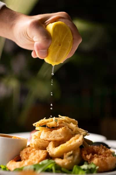 Lemon juice being squeezed over a golden stack of fried vegetables with greens and dipping sauce on the side.