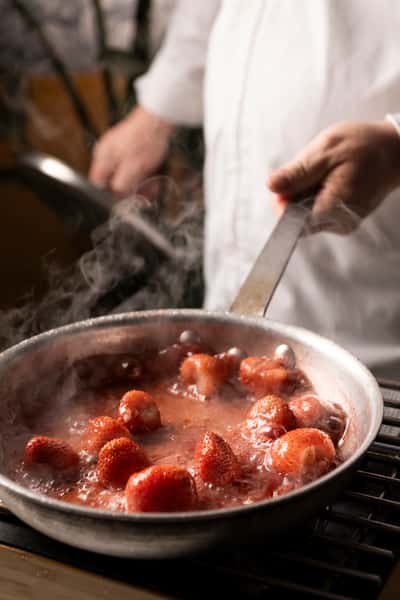Whole strawberries simmering in a bubbling red sauce in a metal pan over heat, with steam rising and a chef stirring in the background.