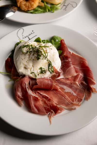 Creamy burrata topped with herbs, served over arugula, cherry tomatoes, and thin slices of prosciutto on a white plate.