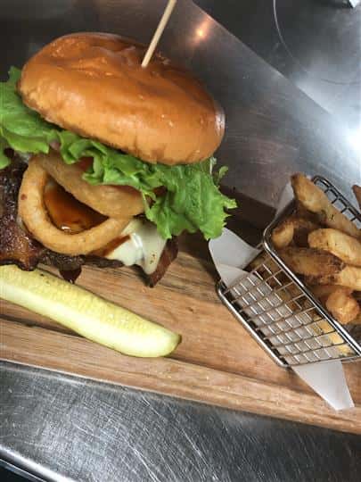 steak sandwich on a bun topped with lettuce and onion rings with a pickle and fries on the side