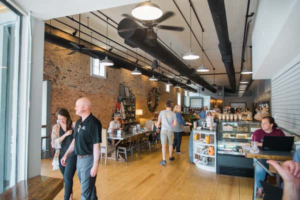 Bright, modern bakery café interior with exposed brick walls, communal seating, pastry display case, and guests enjoying coffee and freshly baked treats.