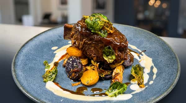 Braised short rib with crispy Brussels sprouts, roasted potatoes, and a cream sauce on a grey plate.