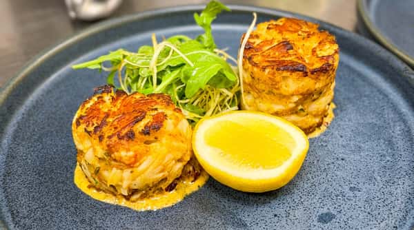 Two crab cakes with a side of mixed greens and a lemon half on a dark gray plate.