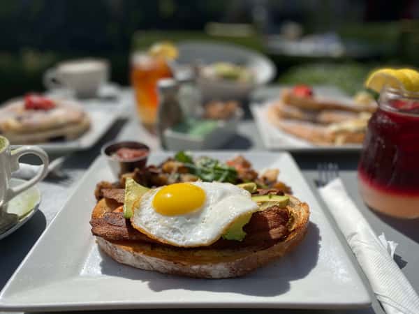 Bright sunny-side up egg and avocado toast served outdoors for brunch, surrounded by drinks and other breakfast dishes.