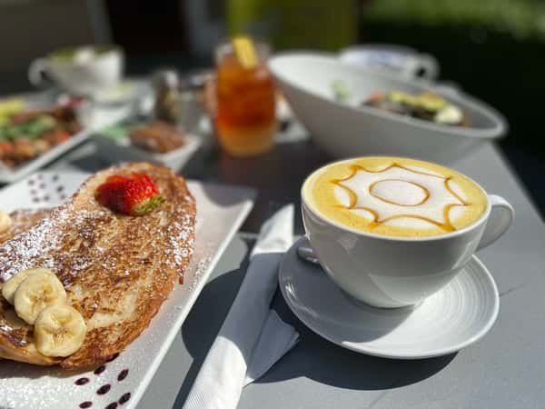 Outdoor breakfast featuring golden French toast with strawberries and bananas, paired with a white mug of caramel latte art.