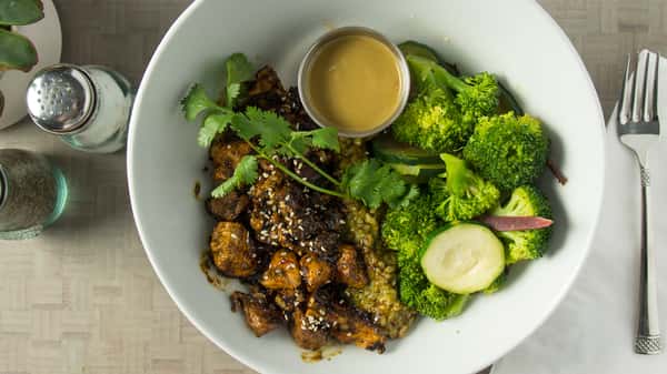 Overhead shot of a white bowl containing teriyaki-glazed chicken, vibrant green broccoli, zucchini, cilantro, and a side of peanut sauce.