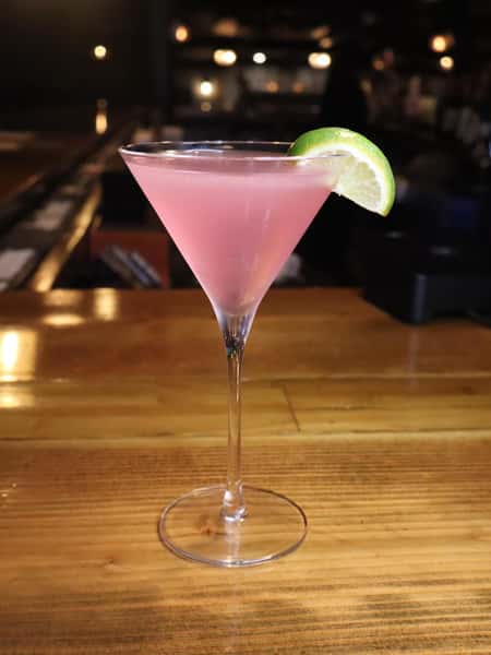 Pink martini cocktail garnished with a fresh lime wedge, served in a classic martini glass on a warm, dimly lit bar.