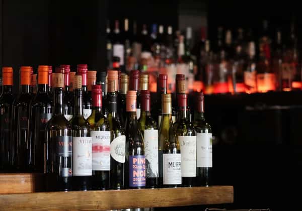 Assorted red and white wine bottles displayed on a wooden bar, with a softly lit back bar creating an upscale restaurant ambiance.