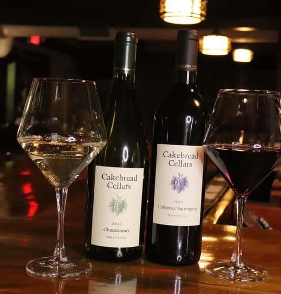 Cakebread Cellars Chardonnay and Cabernet Sauvignon bottles with filled white and red wine glasses on a warmly lit restaurant bar.