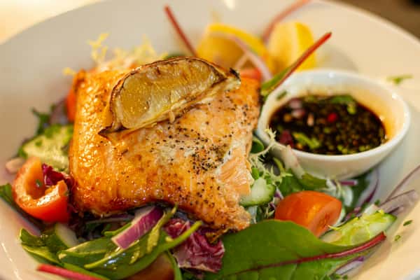 Grilled salmon fillet on a bed of fresh greens with cherry tomatoes, cucumber, red onion, and a charred lemon wedge.