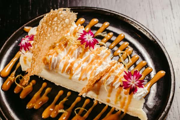 Lemon cream pie topped with whipped cream, caramel drizzle, edible flowers, and sugar crisps on a decorative black plate.