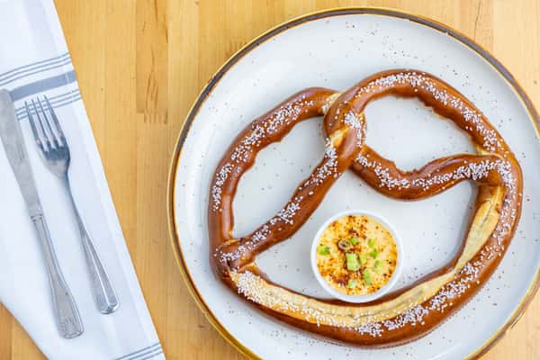 Oversized salted soft pretzel served with a creamy, seasoned beer cheese dipping sauce on a large white plate with gold trim.