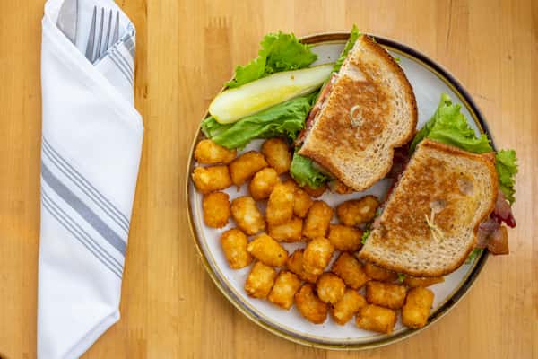Toasty sandwich with lettuce and tomato on whole grain bread, served with golden tater tots and a crisp pickle spear on a white-rimmed plate.