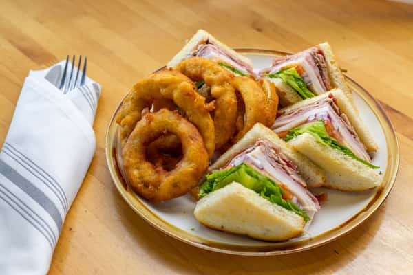 Classic deli-style club sandwich layered with turkey, ham, lettuce, and tomato on toasted white bread, served with crispy golden onion rings on a ceramic plate.
