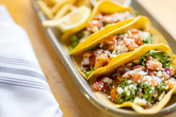 Close-up of fresh chicken street tacos in yellow corn tortillas, packed with lettuce, pico de gallo, and crumbled cheese, served with lemon wedges and fries on the side.
