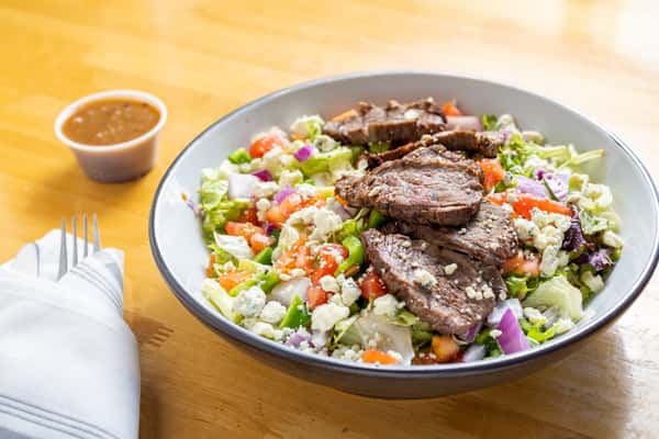 Hearty steak salad with grilled beef slices over a bed of romaine and iceberg lettuce, topped with tomatoes, red onions, blue cheese crumbles, and served with vinaigrette.